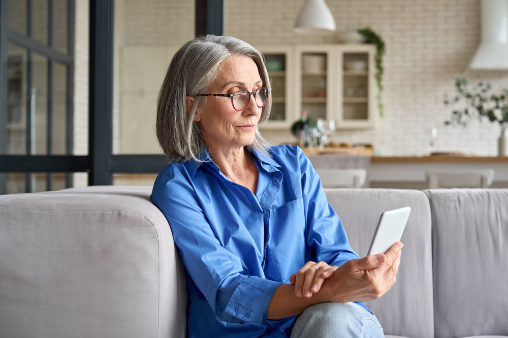 mature woman at home on couch reading cellphone with reading glasses.