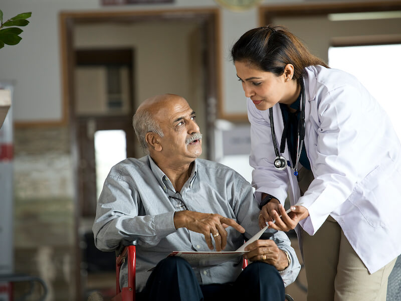 Female doctor communicating with older man who has hearing loss in wheelchair examining reports at the hospital corridor