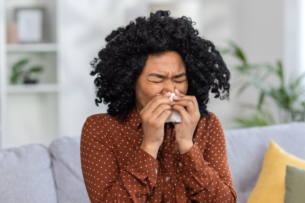 Woman sneezing into tissue at home, showing symptoms of a sinus infection or allergies.