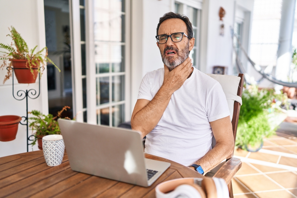 Middle age man using computer laptop at home touching painful neck.