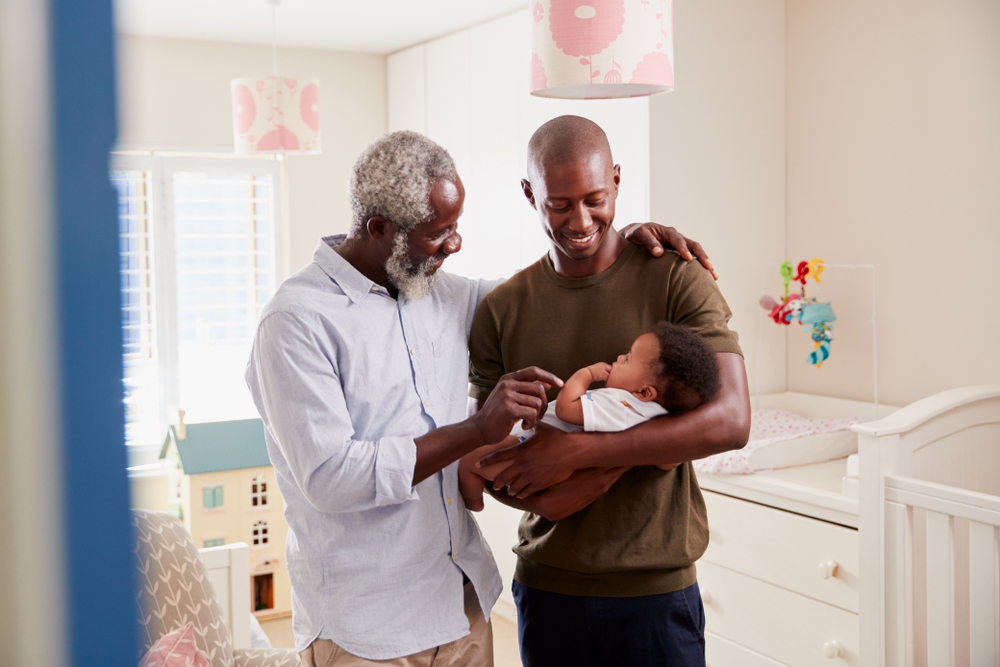 Proud Grandfather With Adult Son Cuddling Baby Grandson In Nursery At Home.