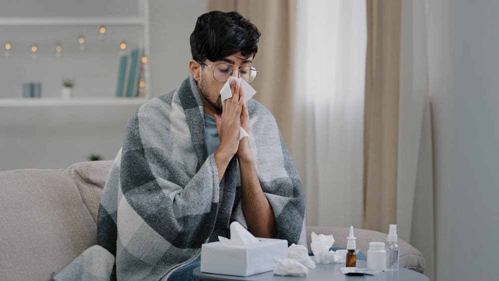 man sitting on couch in blanket blowing nose due to sickness.