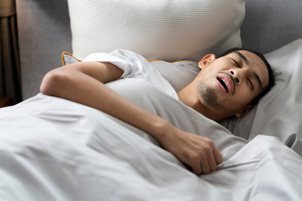 A young bearded man sleeps with his mouth open and makes a loud snore lying down on bed.