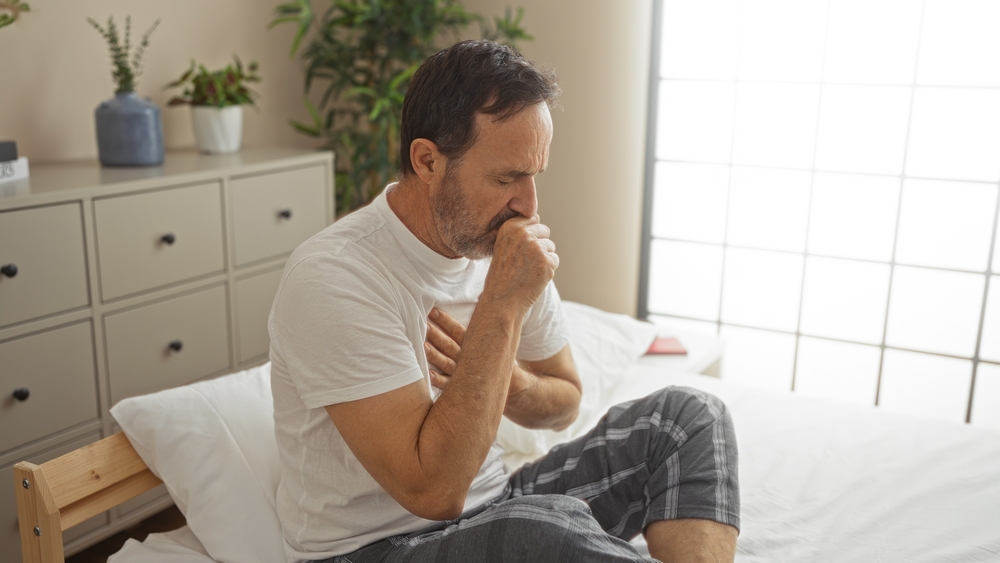 man sitting on bed in bedroom holding his chest while coughing.