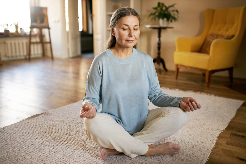 Calm barefoot middle aged female sitting on carpet in half lotus posture.