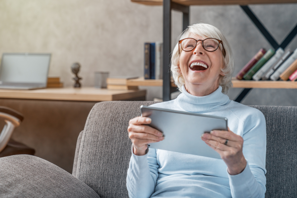 Happy senior woman looking and laughing at her digital tablet on sofa.