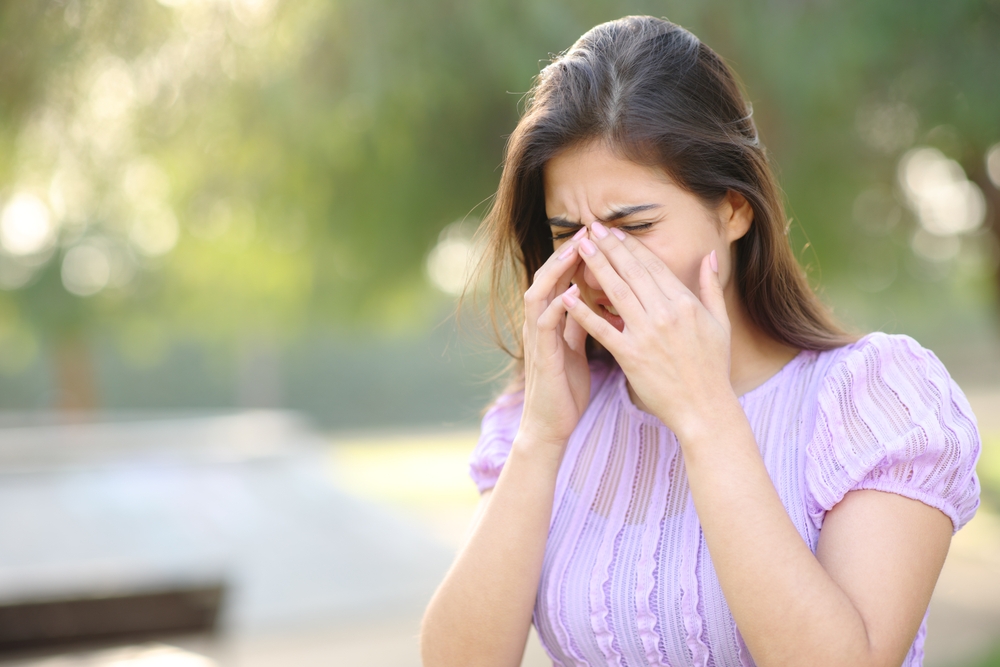 woman suffering from spring allergies, scratching eyes in park outside.